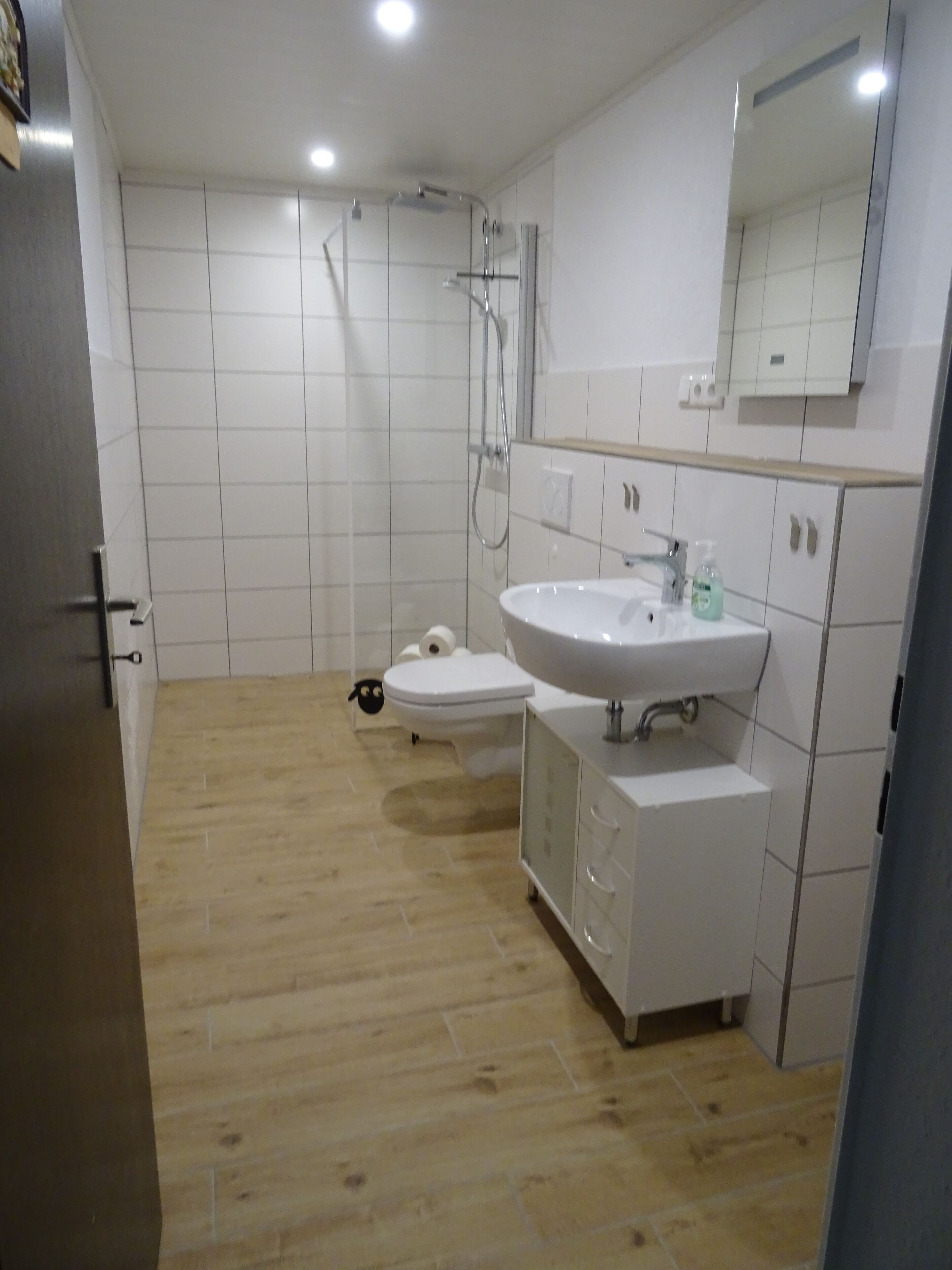 A modern bathroom with a wooden floor, white tiles, a sink, a toilet, and a shower.