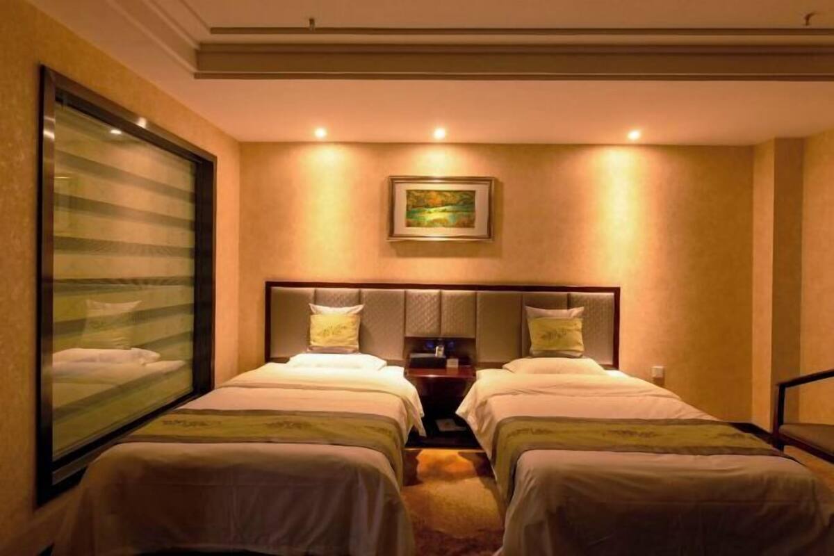 Ein Hotelzimmer mit zwei Betten, einem großen Fenster mit Jalousien, einem Bild an der Wand und einem kleinen Tisch zwischen den Betten.
