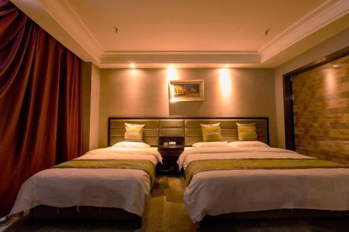 Ein Hotelzimmer mit zwei Betten, einem an der Bettseite angebrachten Nachtbaud mit Uhr, einem Bild an der Wand und einem großen Fenster mit Vorhängen.