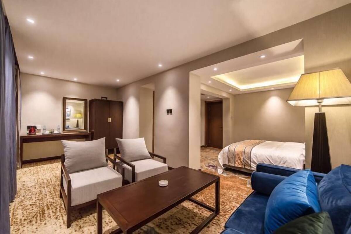 Habitación de hotel con una cama, un sofá azul, dos sillones, una mesa de centro y un escritorio con una lámpara.