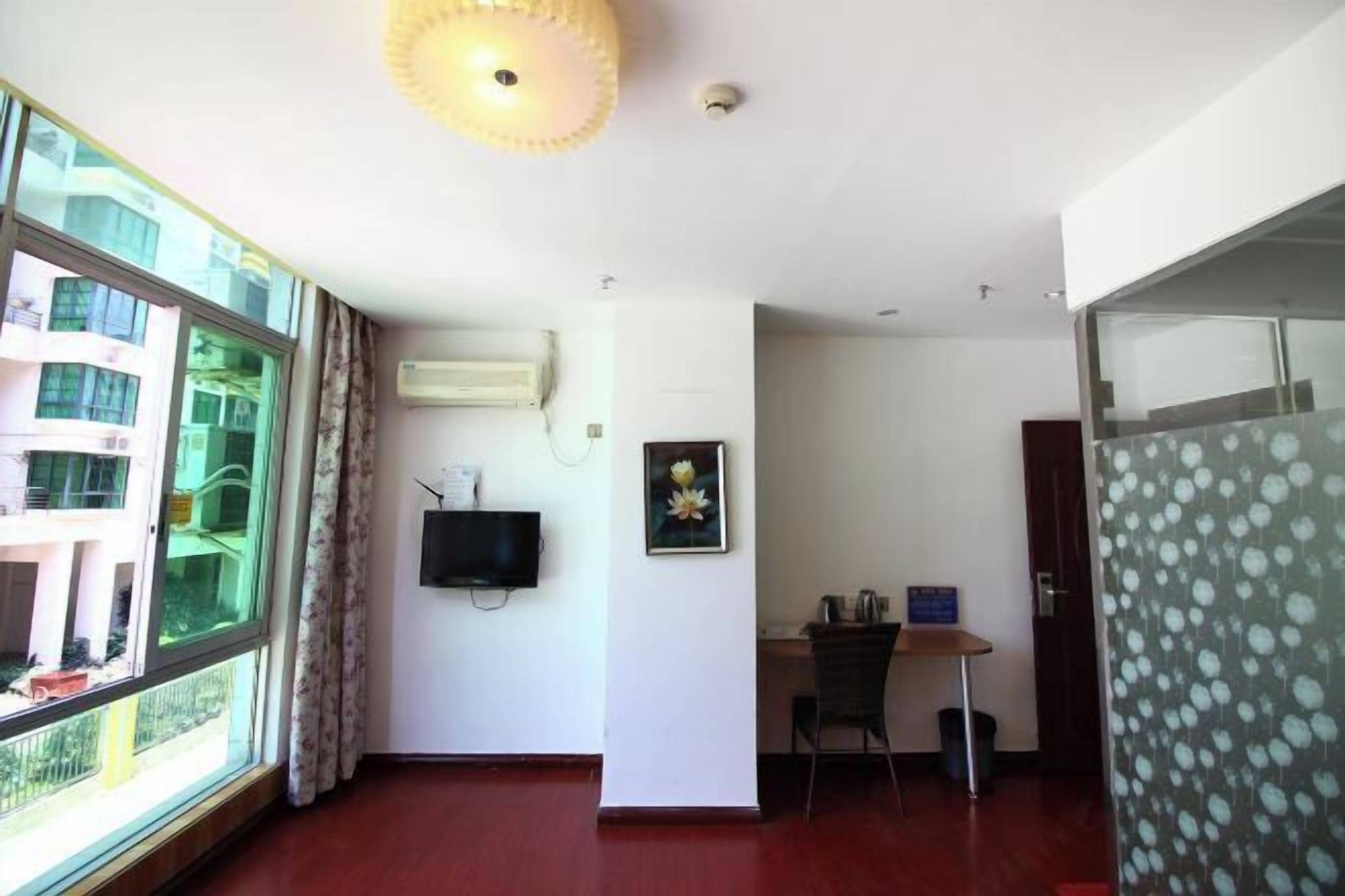 Una habitación con un ventanal grande, un televisor de pantalla plana montado en la pared, un escritorio con una silla y una puerta que da a otra habitación.