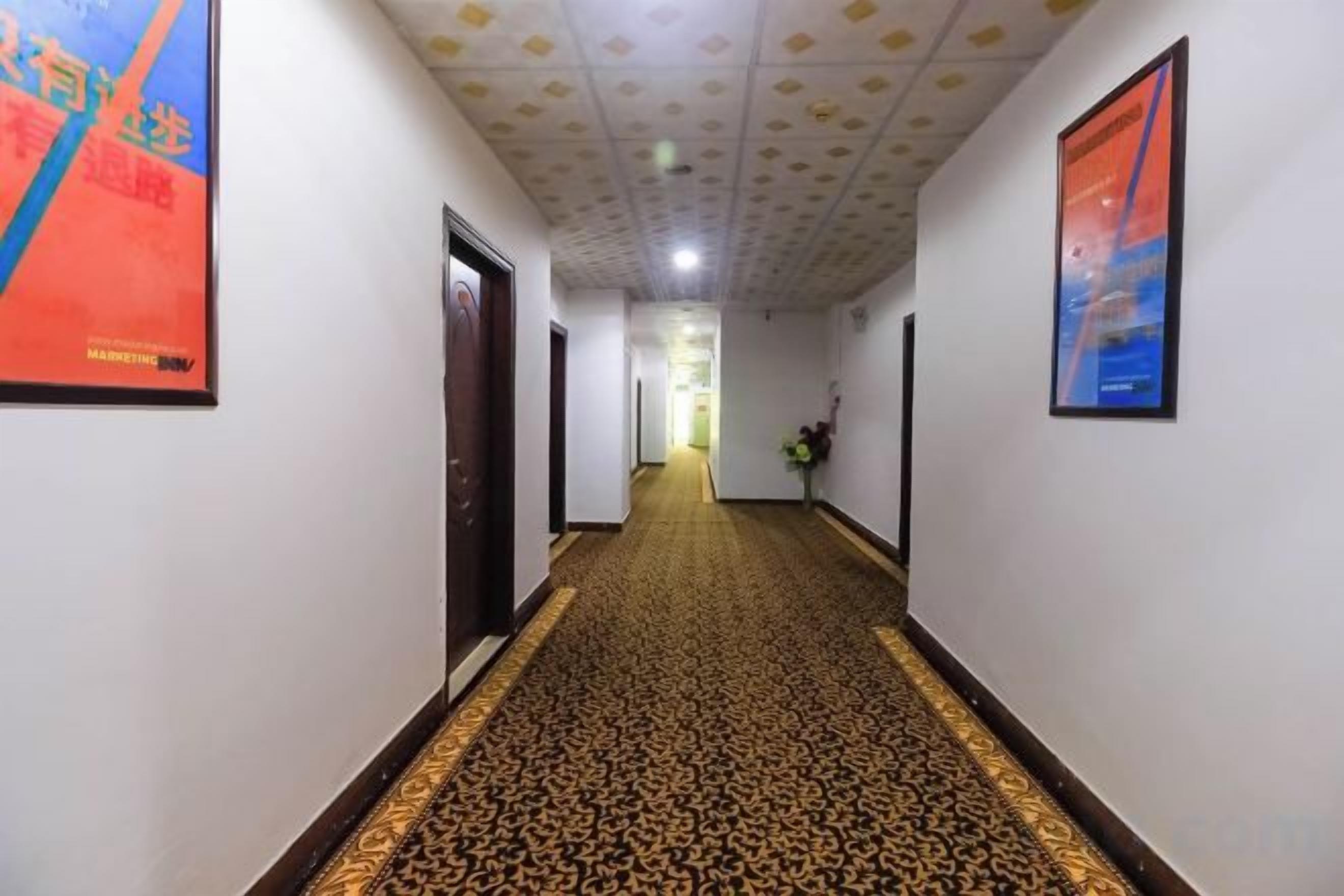 A hotel hallway with patterned carpet, framed artwork on the walls, and doors leading to rooms.