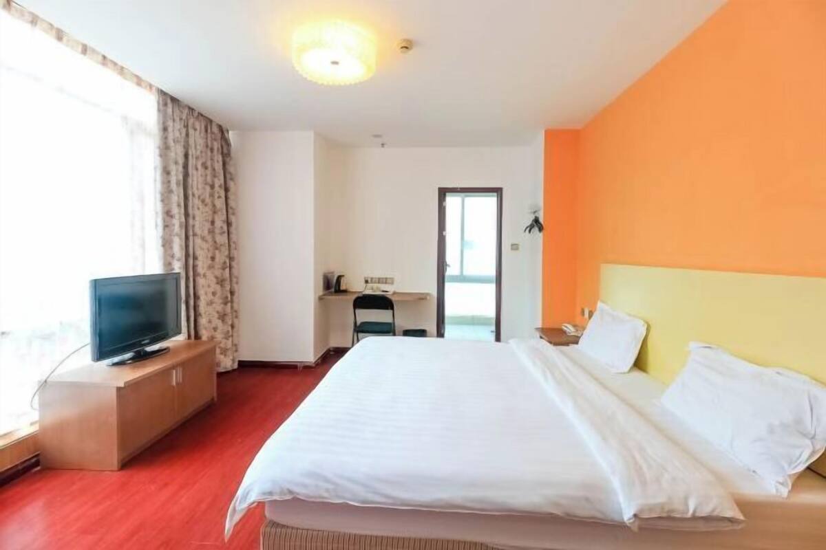 Habitación de hotel con cama, televisor sobre un mueble de madera, escritorio con silla y vista a través de una puerta.