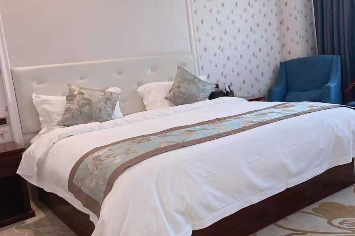 A hotel room with a large bed, a headboard, a blue armchair, and a patterned curtain.