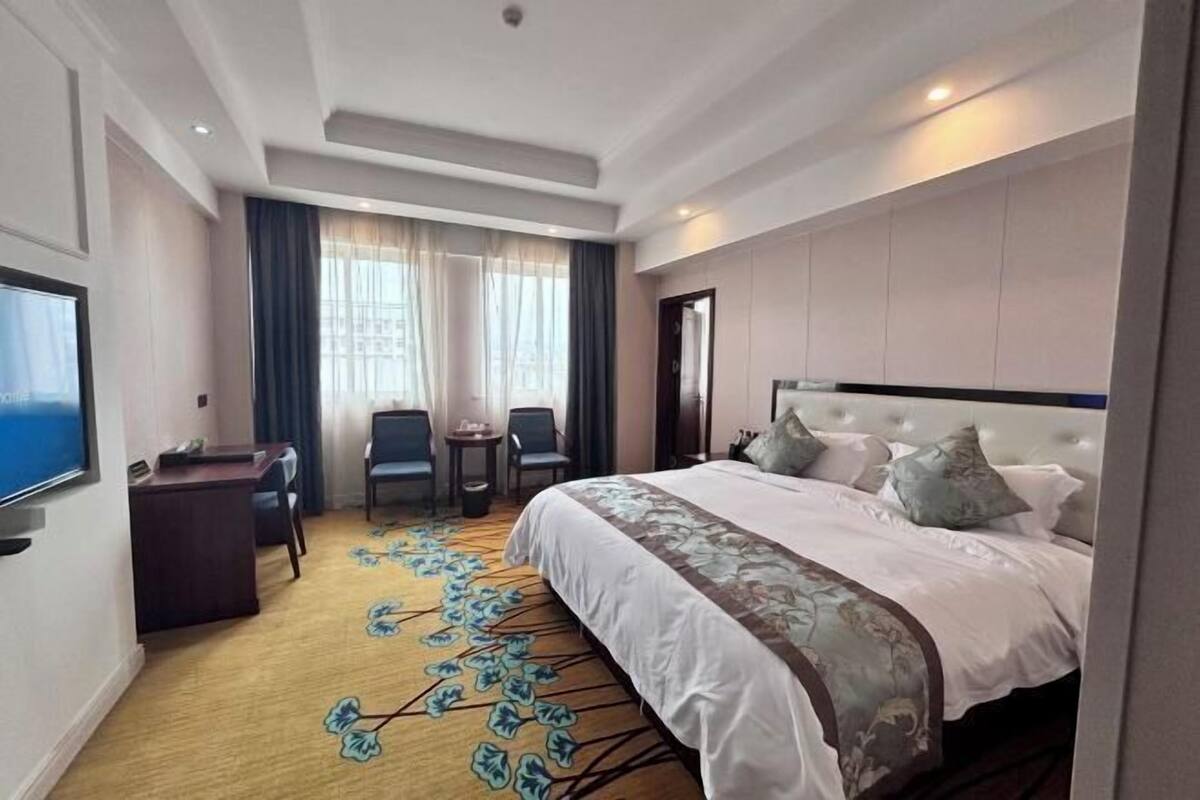 A hotel room with a large bed, a desk, a TV, and a view of the city through the window.