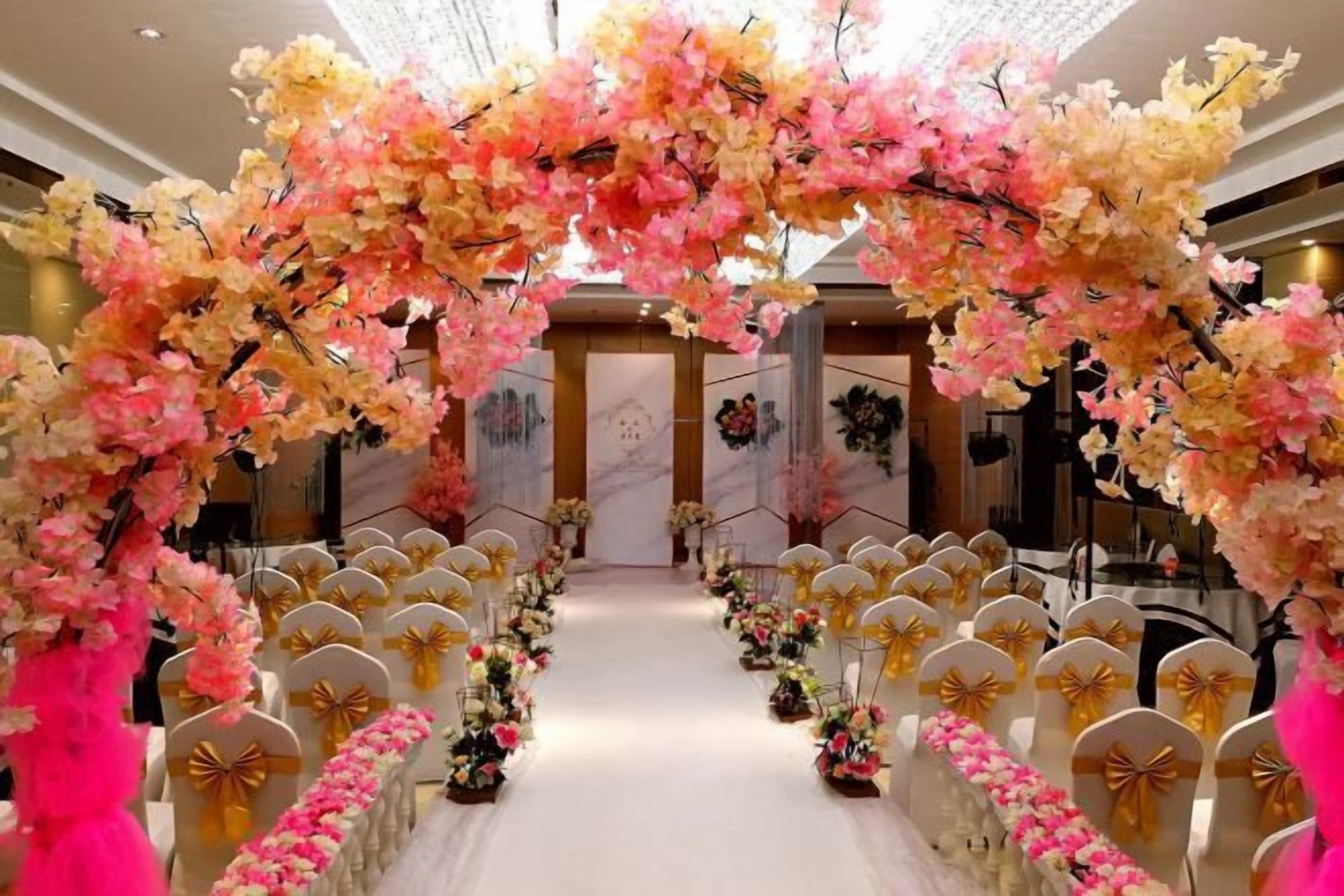 Interior de hotel con un arco floral, filas de sillas con lazos dorados y un suelo de mármol.