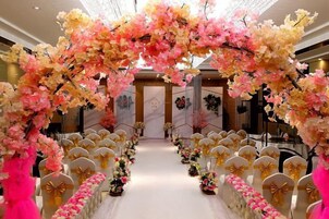 Interior de hotel con un arco floral, filas de sillas con lazos dorados y un suelo de mármol.