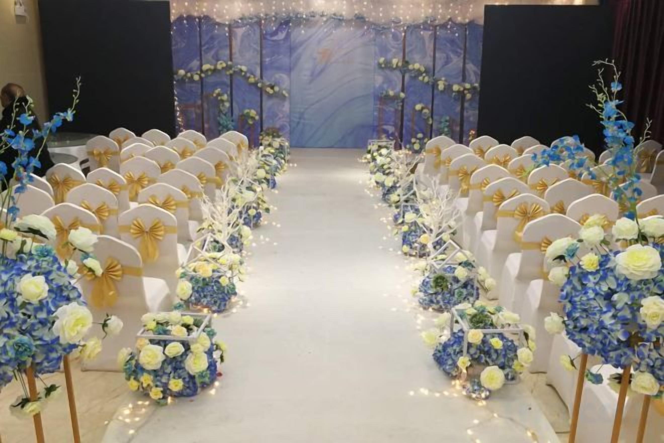 A venue with rows of chairs and floral decorations, a stage with a backdrop, and a carpeted floor.