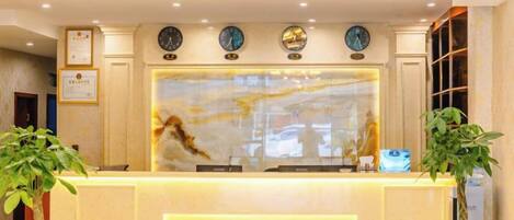A hotel reception area with a marble counter, clocks, and potted plants.