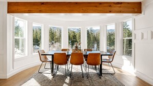 Comedor con una mesa grande y seis sillas, ventanas amplias con vistas a árboles y una viga de madera en el techo.