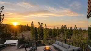 Una terraza con un pozo de fuego, mobiliario exterior y vistas a una puesta de sol sobre un bosque.