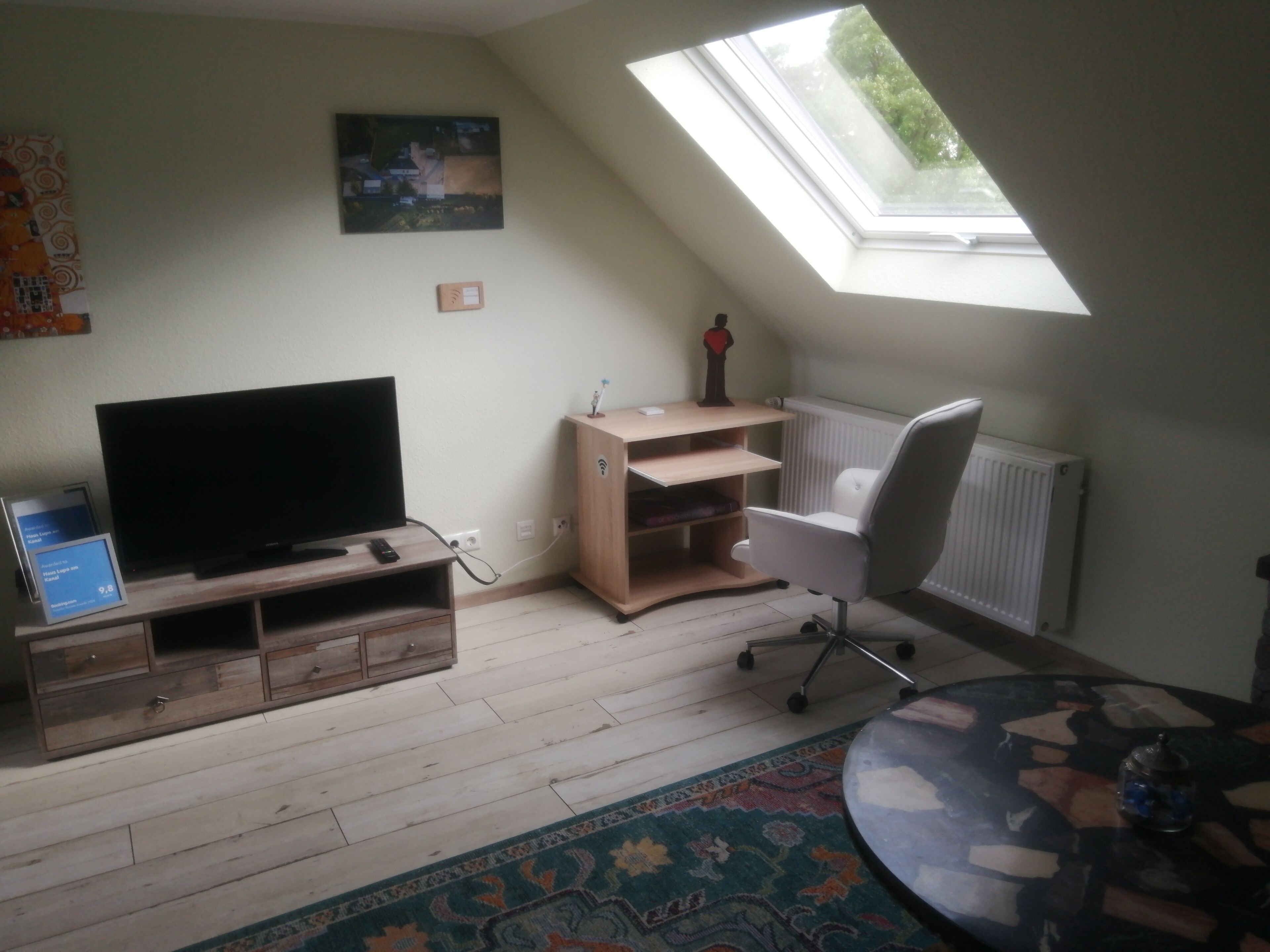 Ein Zimmer mit Fernseher auf einem Holzgestell, einem Schreibtisch mit Stuhl, einer Heizung und einem Oberlicht.