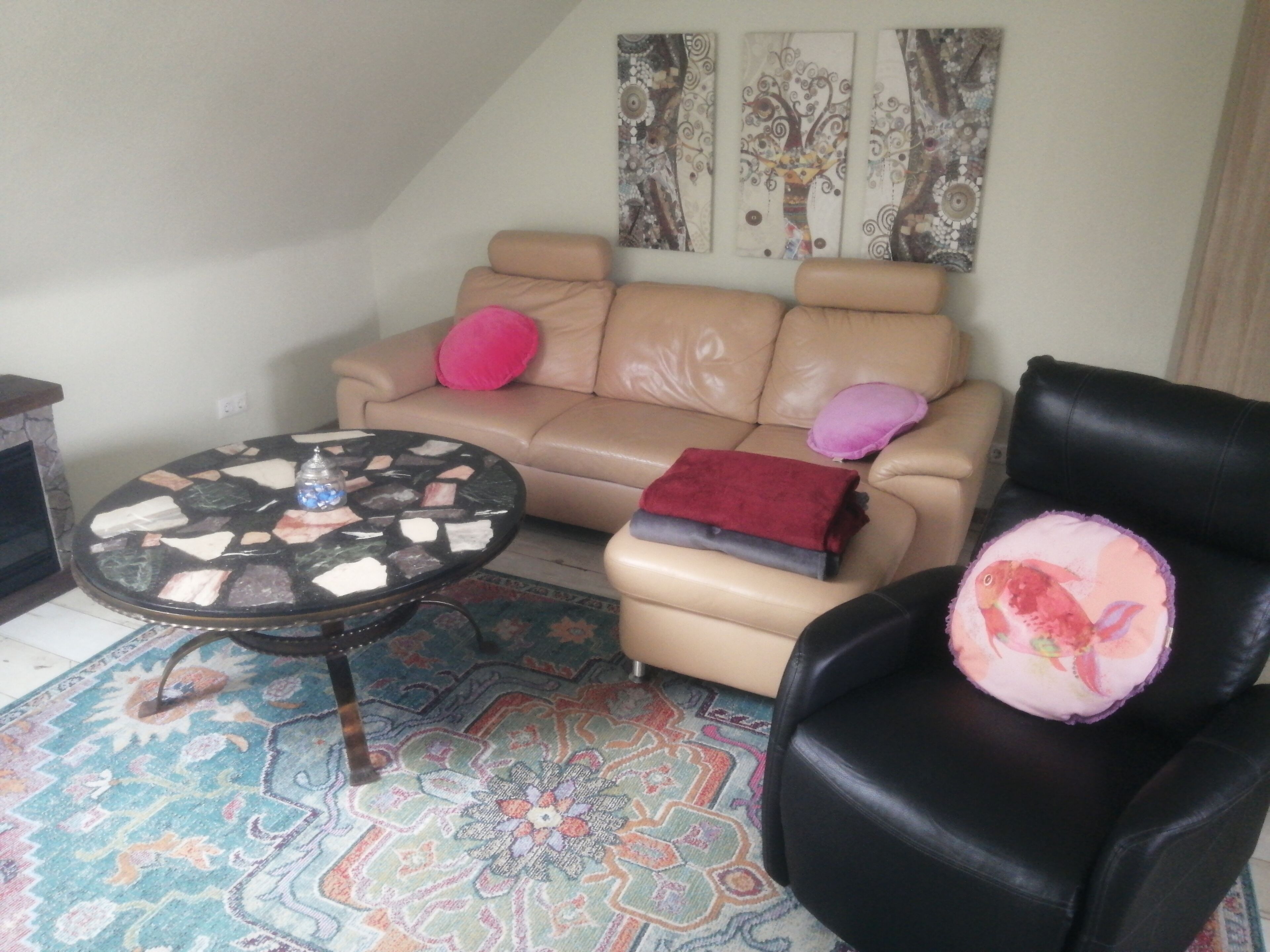 A living room with a beige sofa, a black leather chair, a round coffee table with a mosaic top, and a patterned rug.