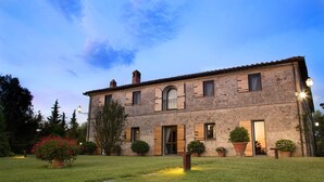 Un edificio de piedra de dos pisos con un jardín delantero, que incluye una planta grande en maceta y un banco.
