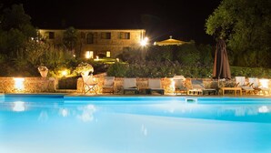 Área de piscina nocturna con sillones reclinables y un edificio al fondo.