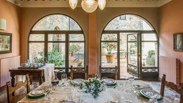Un comedor con una mesa lista para comer, ventanas arqueadas grandes y una pared de piedra visible a través de las ventanas.