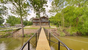 Een houten brug die leidt naar een twee verdiepingen tellend huis met een balkon, omgeven door groen en een rivier.