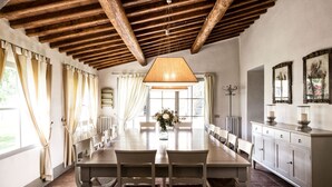 A dining room with a long table, white chairs, a hanging light fixture, and a view of the outdoors through large windows. - Wonderful Villa in the heart of Chianti (Greve in Chianti)
