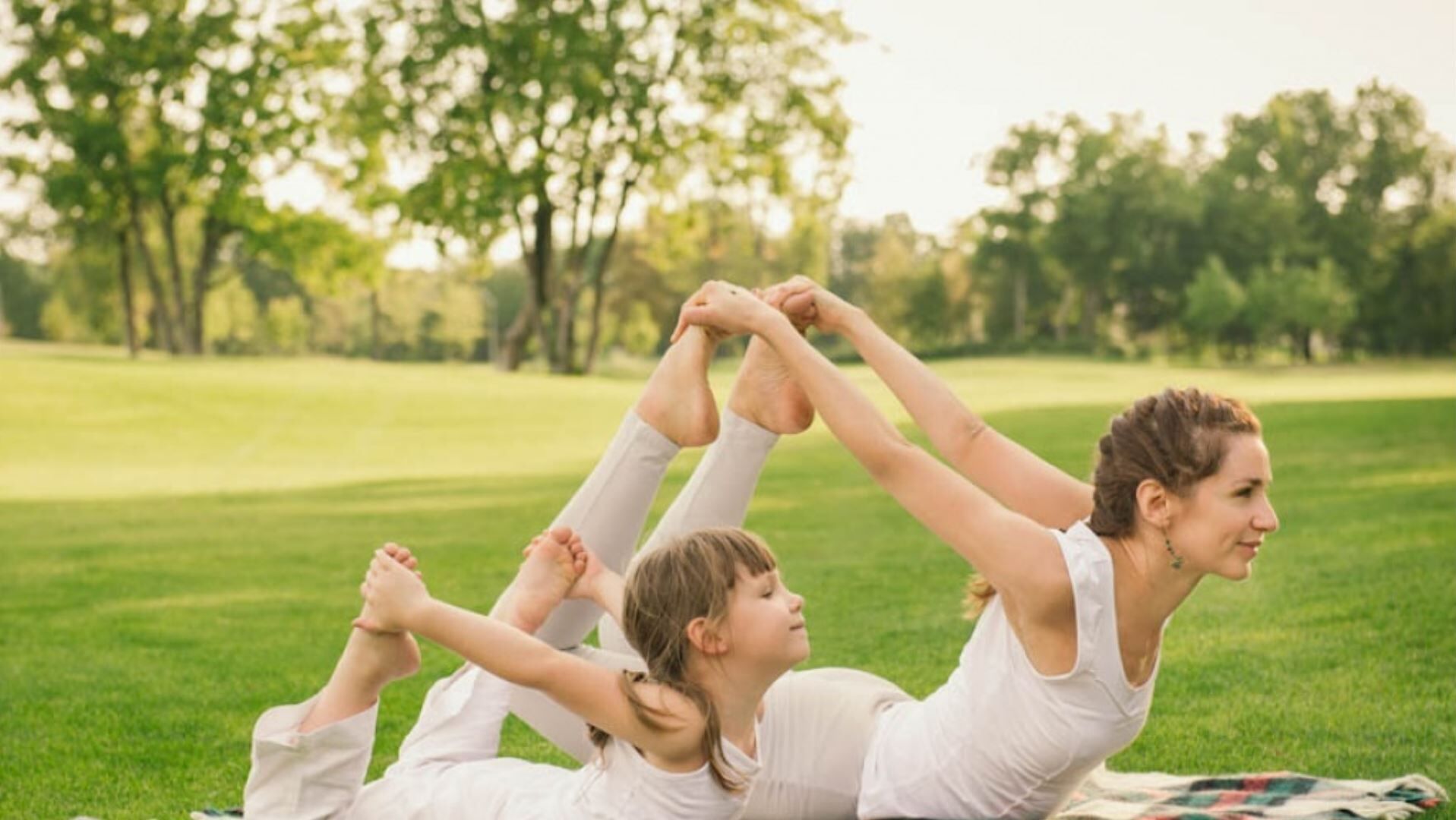 Three people in white performing a yoga pose on a grassy field with trees in the background.