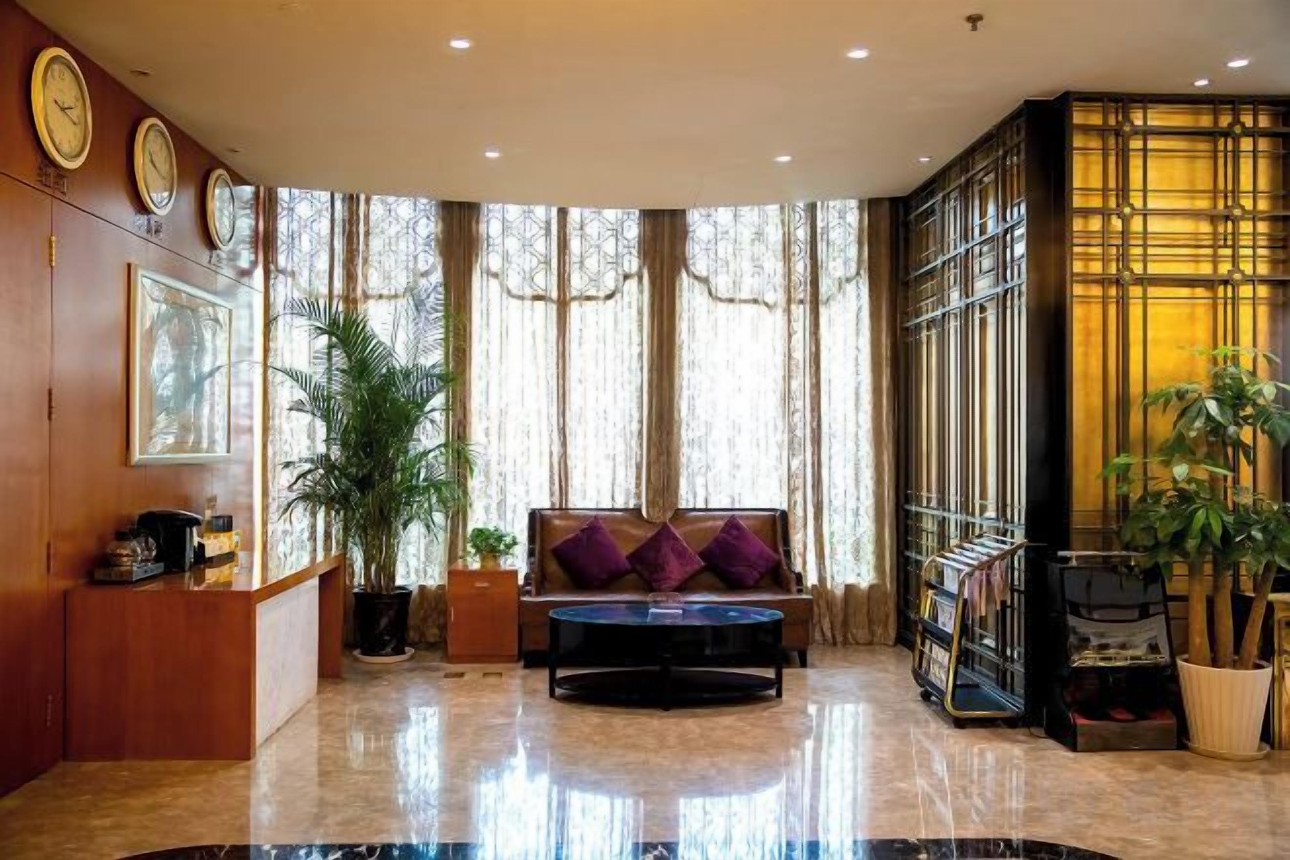 A hotel lobby with a reception desk, seating area, large windows, and decorative plants.