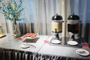 Een buffetopstelling met twee koffieautomaten, een bord met watermeloen en een vaas met kunstbloemen.