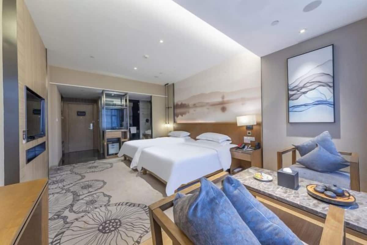 Ein Hotelzimmer mit zwei Betten, einer Sitzecke mit Tisch, einem an der Wand hängenden Kunstwerk und einem durch eine offene Tür einsehbaren Badezimmer.