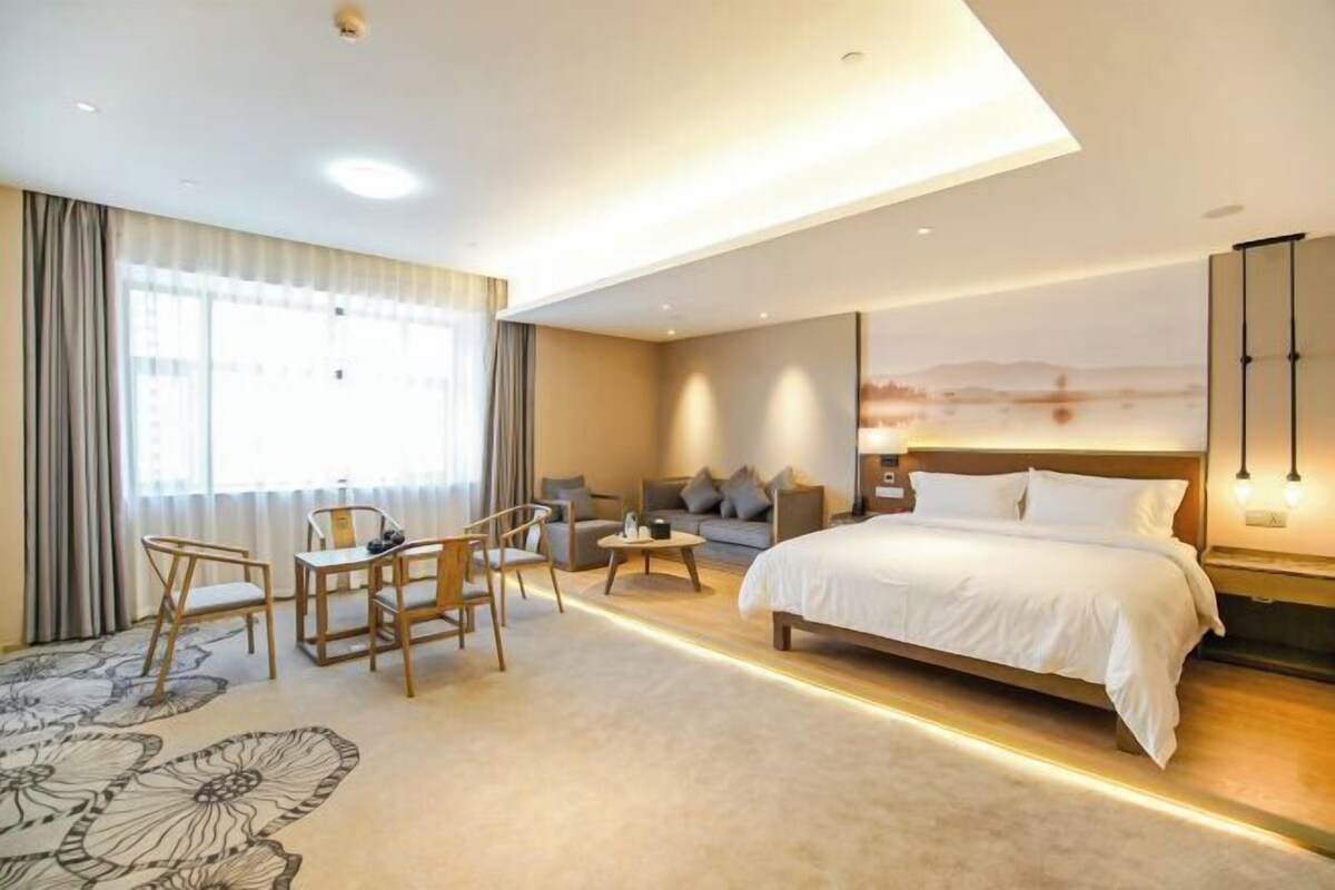 A modern hotel room with a large bed, a sofa, a dining area with chairs, and a view of the cityscape through the window.