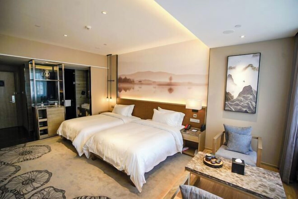 A hotel room with a large bed, a desk with a lamp, a painting of mountains, and a view of a lake.