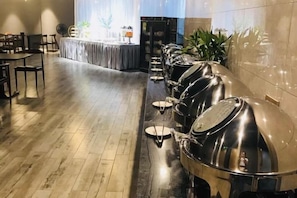 A long buffet line with stainless steel serving stations and a table set for dining in the background.