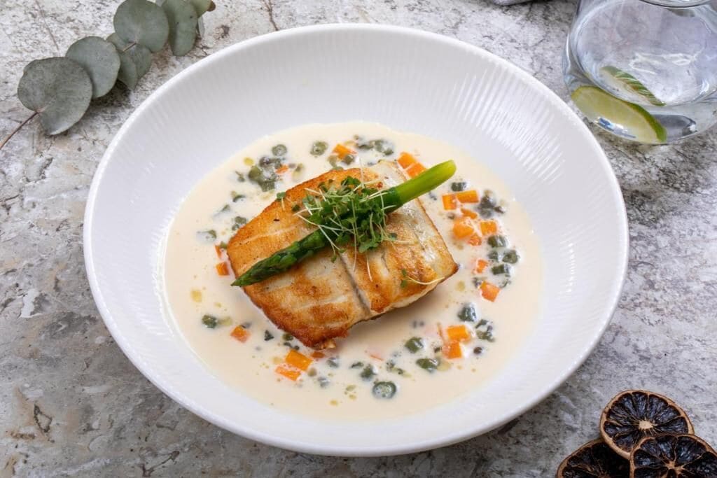 Ein weißer Teller mit Fisch, Spargel und cremiger Soße auf einer Marmorplatte.