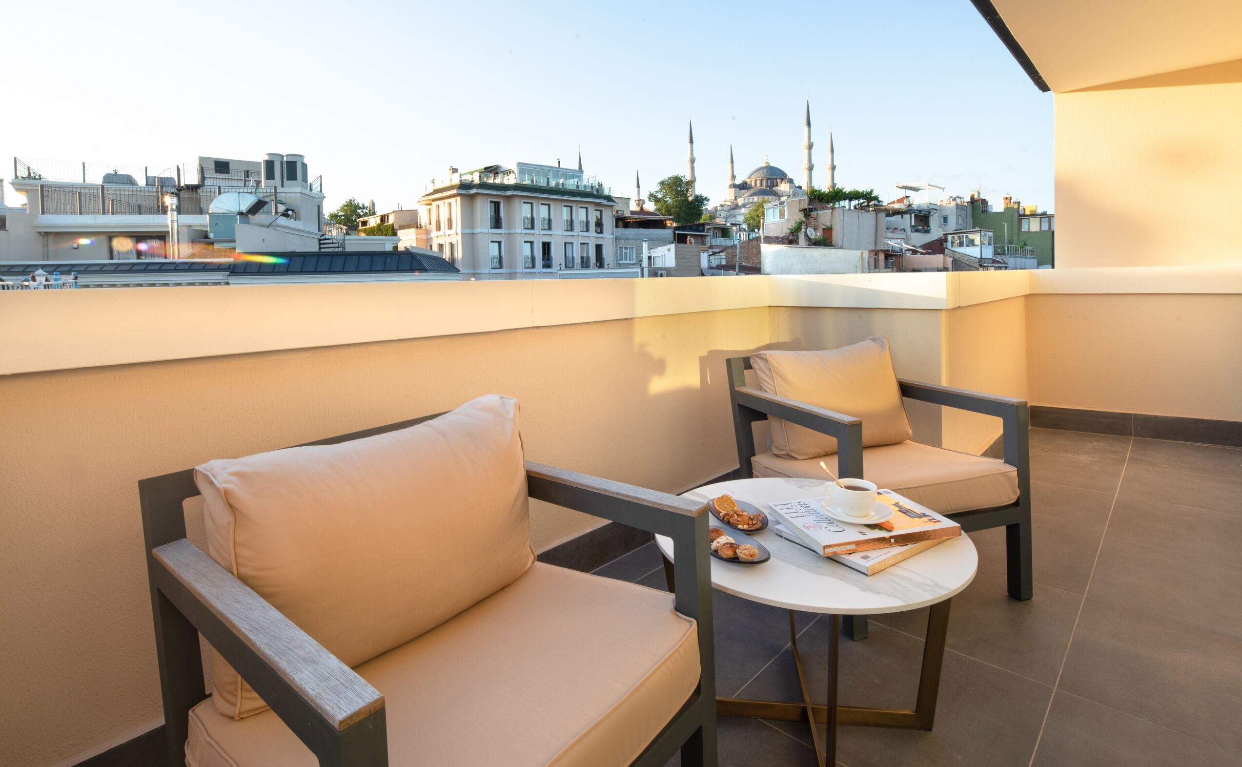 Un balcon avec deux fauteuils beiges dotés de coussins, une petite table ronde sur laquelle se trouvent un livre et une tasse, et une vue sur la ville en arrière-plan.