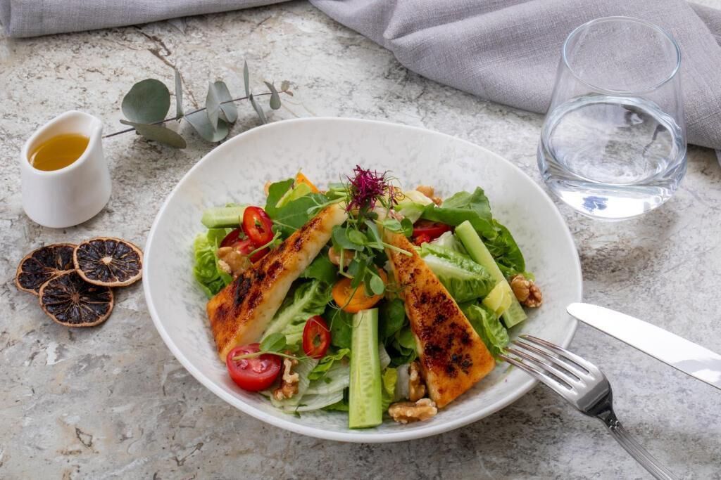 A plate of grilled chicken and salad with a small bowl of dressing and a glass of water on a marble surface.