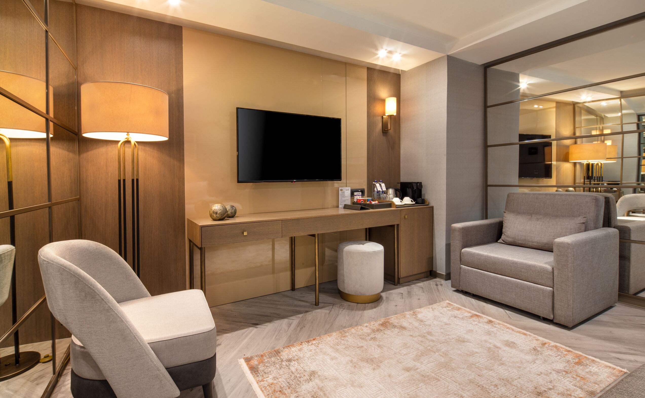 A modern hotel room with a flat-screen TV, a desk with a lamp, two armchairs, and a large mirror.