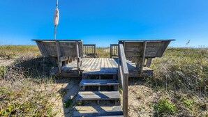 Una pasarela de madera sobre la playa con escalones que dan a una zona arenosa, rodeada de vegetación.