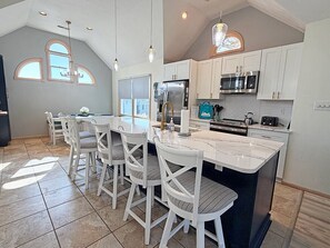 A modern kitchen with white cabinetry, a central island, and a dining area with white chairs. - 🏖️Anchored Inn - updated 4BR/3BA Oceanside home w/Private Pool 🏖️ (Corolla)