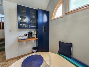 Une cuisine compacte avec des meubles bleus, un coin café et un petit espace détente agrémenté d’un coussin.