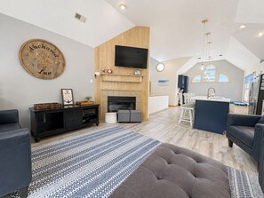 A modern living room with a fireplace, a flat-screen TV mounted above it, and a dining area with a kitchen in the background.