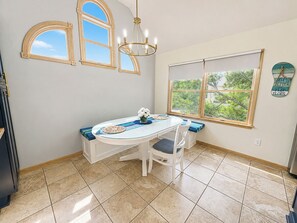 A dining area with a white table, two chairs, a chandelier, and a window with a view of greenery. - 🏖️Anchored Inn - updated 4BR/3BA Oceanside home w/Private Pool 🏖️ (Corolla)