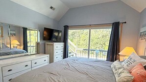 A bedroom with a large bed, a TV, a dresser, and a balcony with a view of trees. - 🏖️Anchored Inn - updated 4BR/3BA Oceanside home w/Private Pool 🏖️ (Corolla)