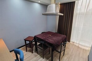 Um quarto pequeno com uma mesa e cadeiras, um respiradouro no teto, uma janela com cortinas e um abajur.