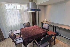 Un coin repas avec une table recouverte d’une nappe sombre, six chaises, une hotte de cuisine moderne, une fenêtre avec des rideaux légers et un petit espace cuisine comprenant une machine à café.
