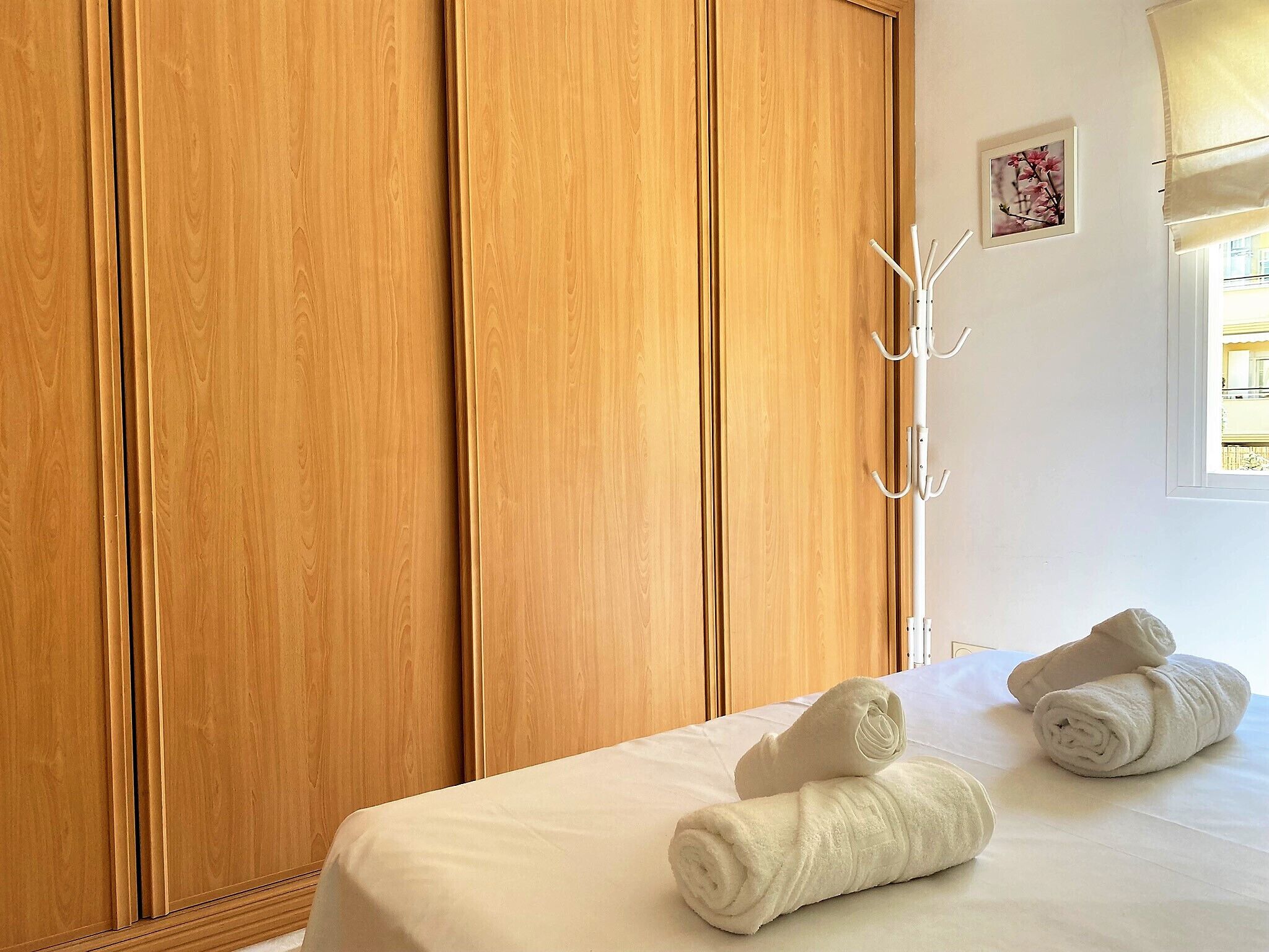 Una camera d'albergo con ante di armadio in legno, un letto bianco con due asciugamani arrotolati, una parete bianca e una finestra con vista.