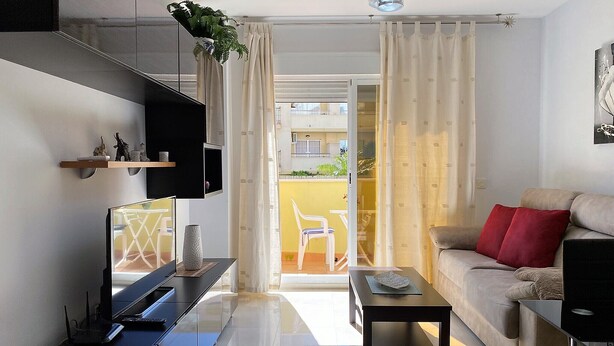A modern living room with a sofa, a coffee table, a TV, and a balcony with a view of the outside.