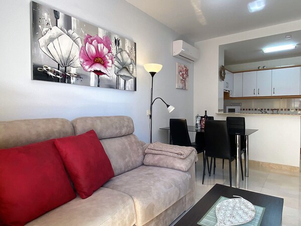 A living room with a beige sofa, a red pillow, a floor lamp, and a dining area with a table and chairs.