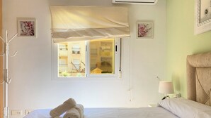 A hotel room with a bed, two rolled towels, a window with a view of the outside, a bedside lamp, and a wall-mounted air conditioner.