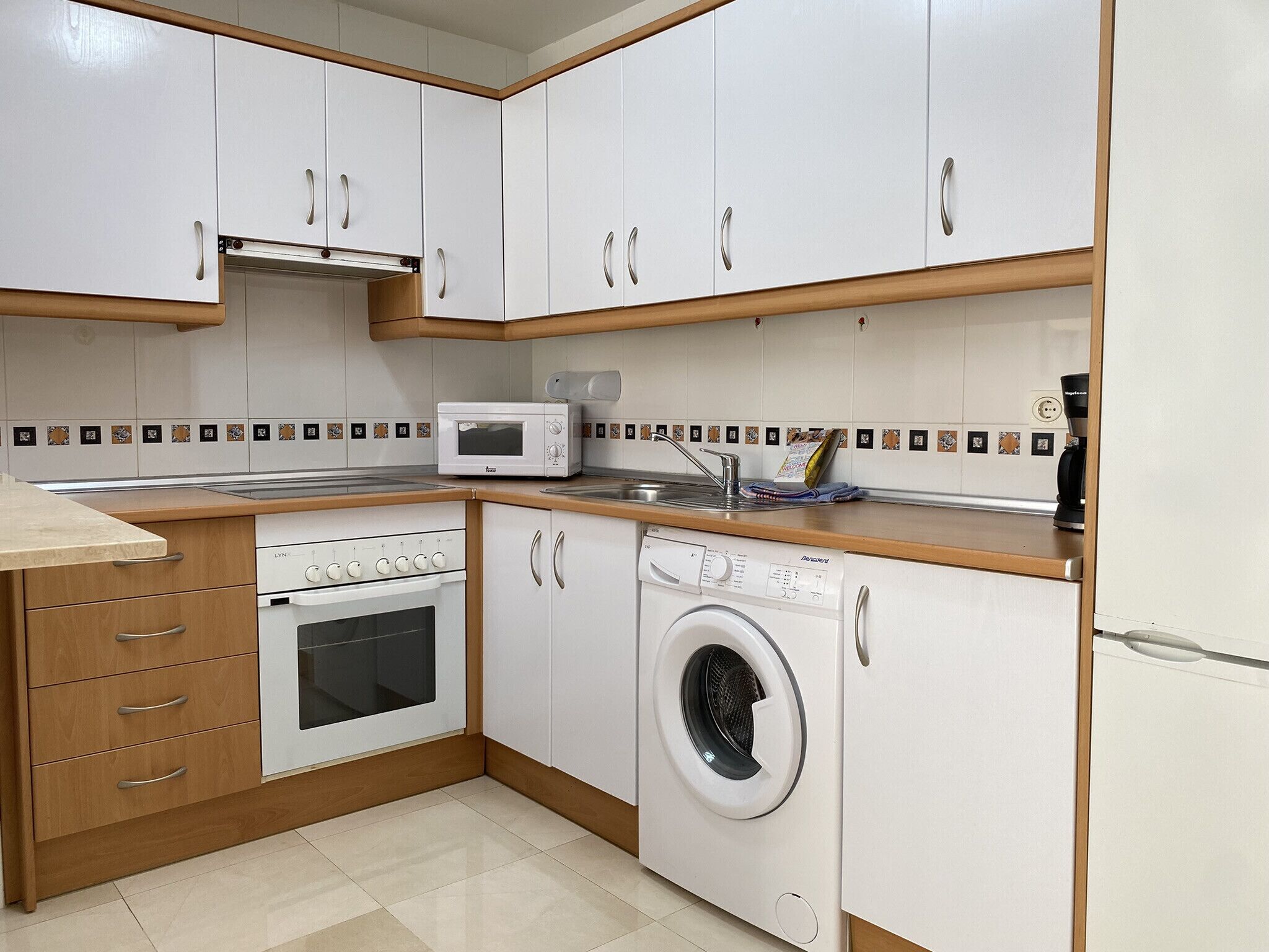 Een compacte keuken met witte kastjes, een magnetron, een oven, een wasmachine en een koffiezetapparaat.