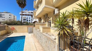 A swimming pool with a stone wall and a metal fence, surrounded by a building with balconies and potted plants.
