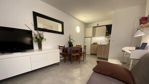 Una sala de estar moderna con televisor de pantalla plana, una zona de comedor con sillas de madera, una cocina con armarios de madera y un sofá blanco.