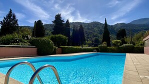 Una piscina azul cristalina con barandillas metálicas, rodeada por un jardín ajardinado y vista a una ladera boscosa bajo un sol brillante.