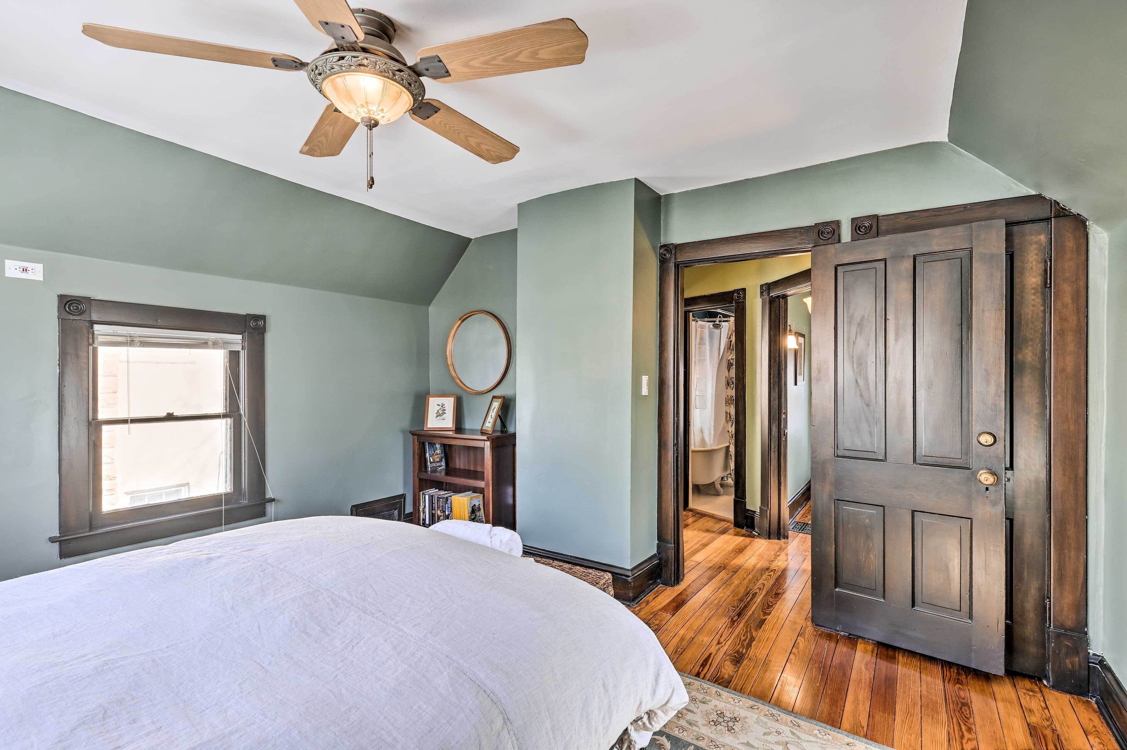 Une chambre à coucher comprenant un lit, un ventilateur de plafond, une fenêtre, une porte en bois et un couloir menant à une autre pièce.